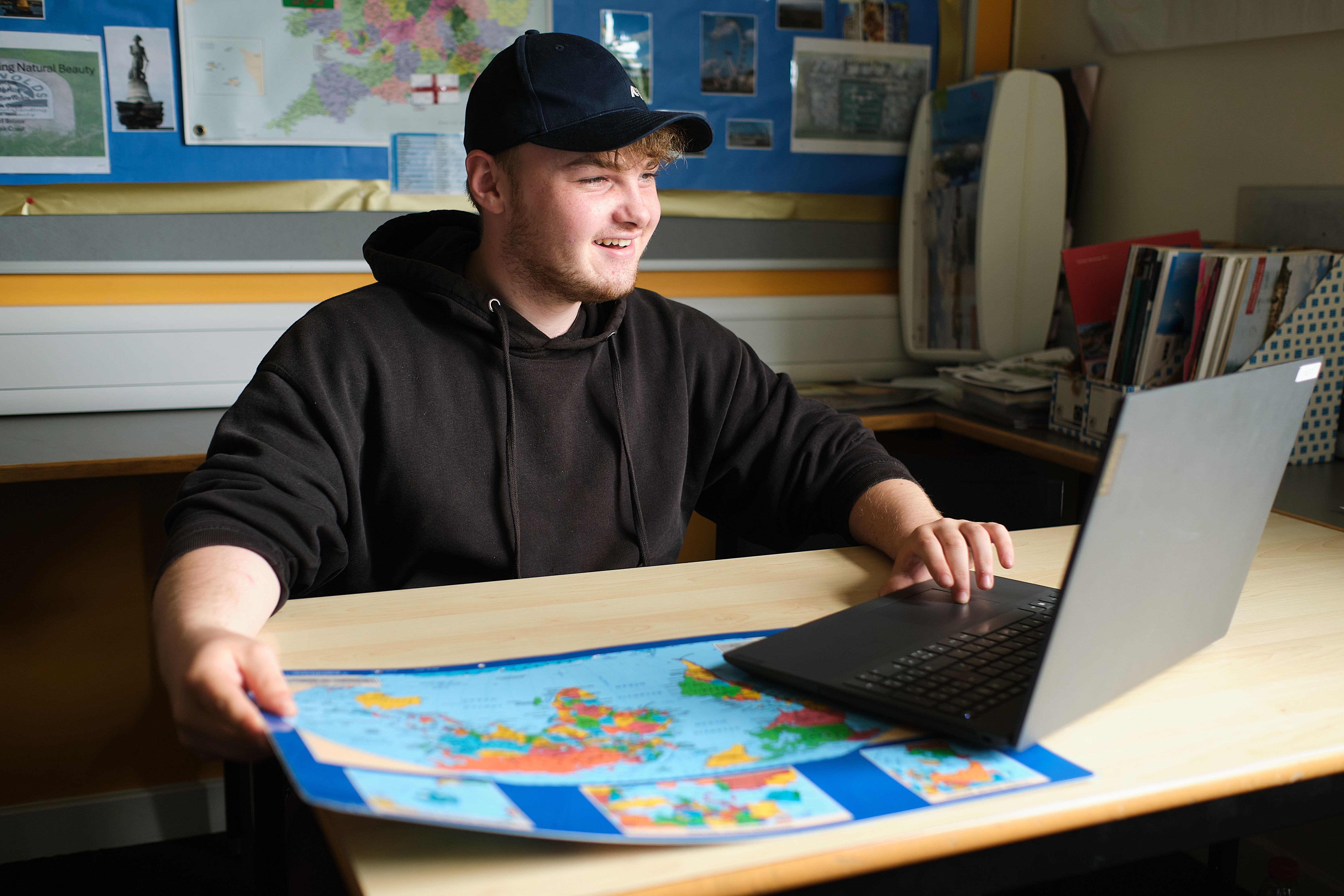 A student smiles while working on a laptop and holding a map in a classroom.