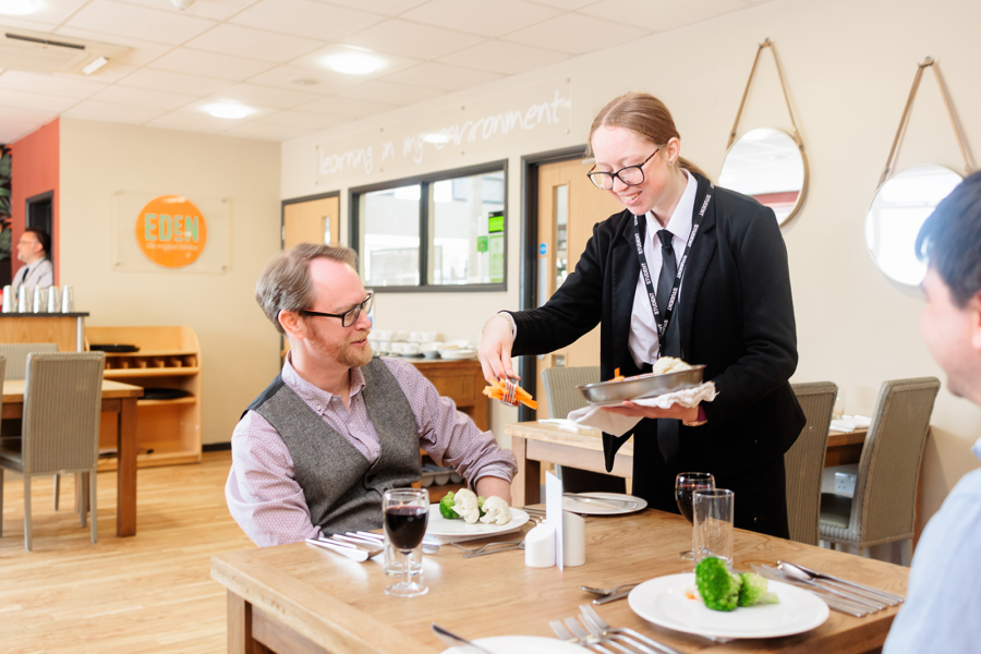 Student waiting staff serving guests at a restaurant table.