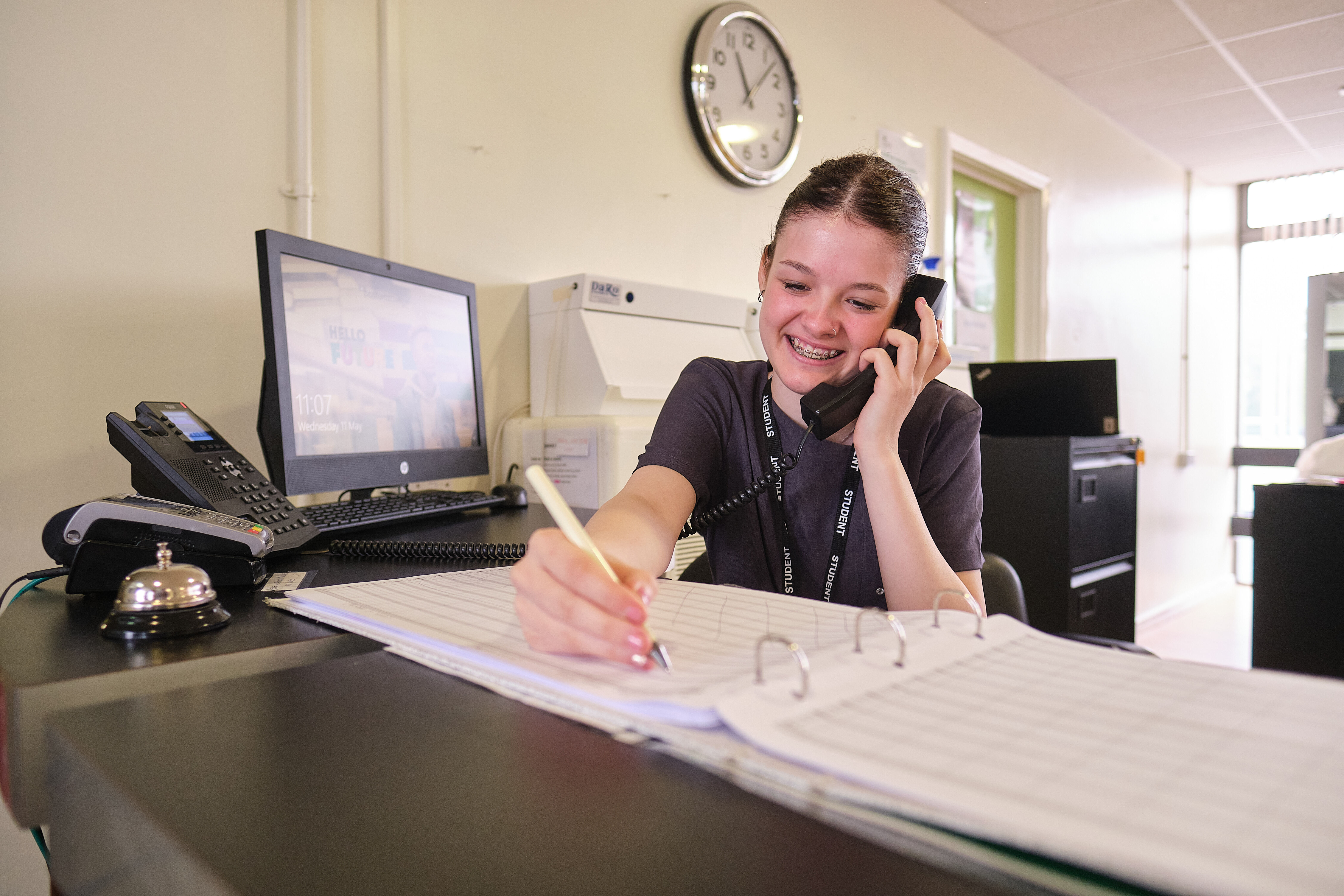 A student taking a call and writing in a book at a reception desk.