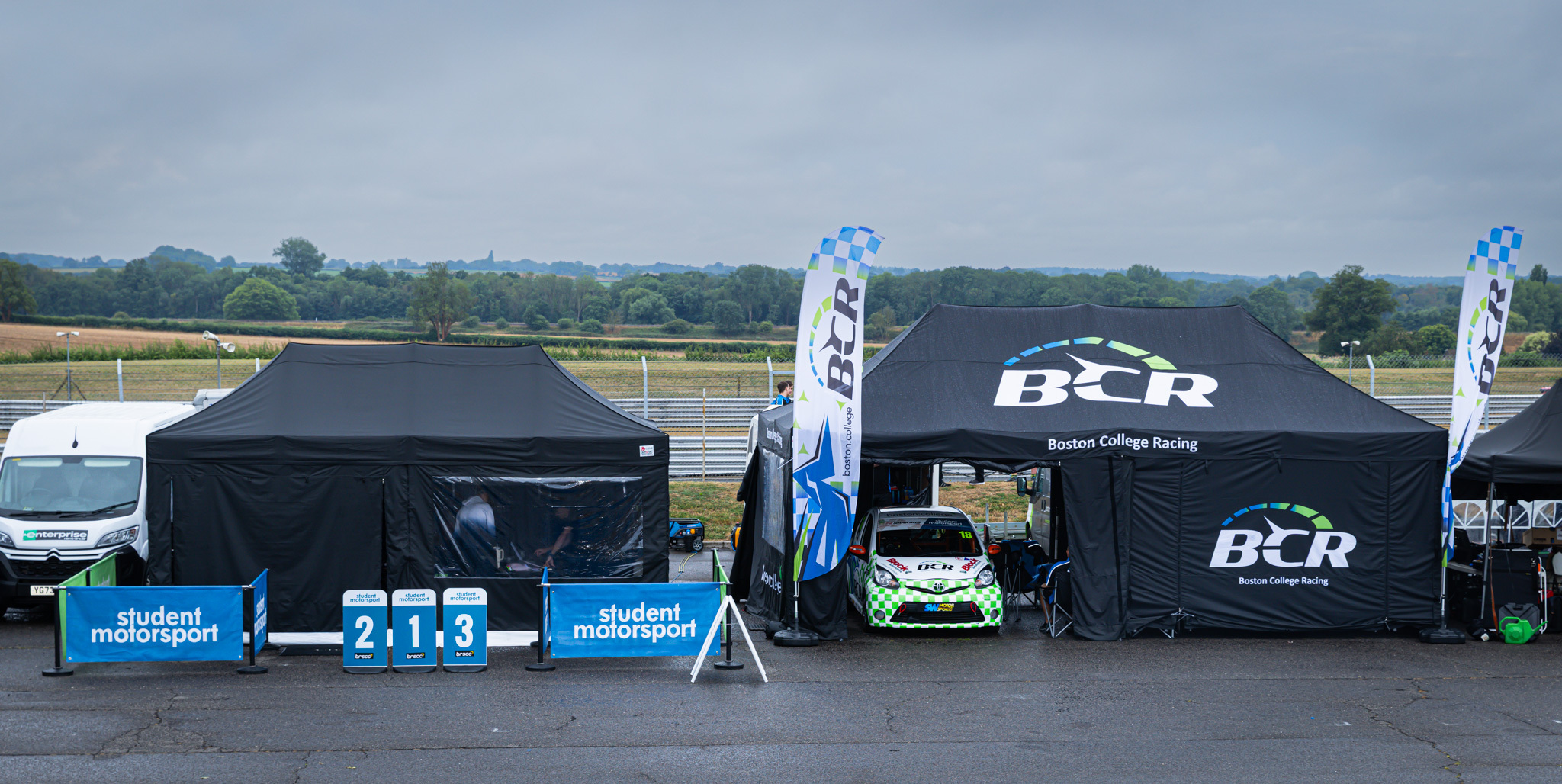 Boston College Racing cars under a gazebo at a wet and grey racetrack.