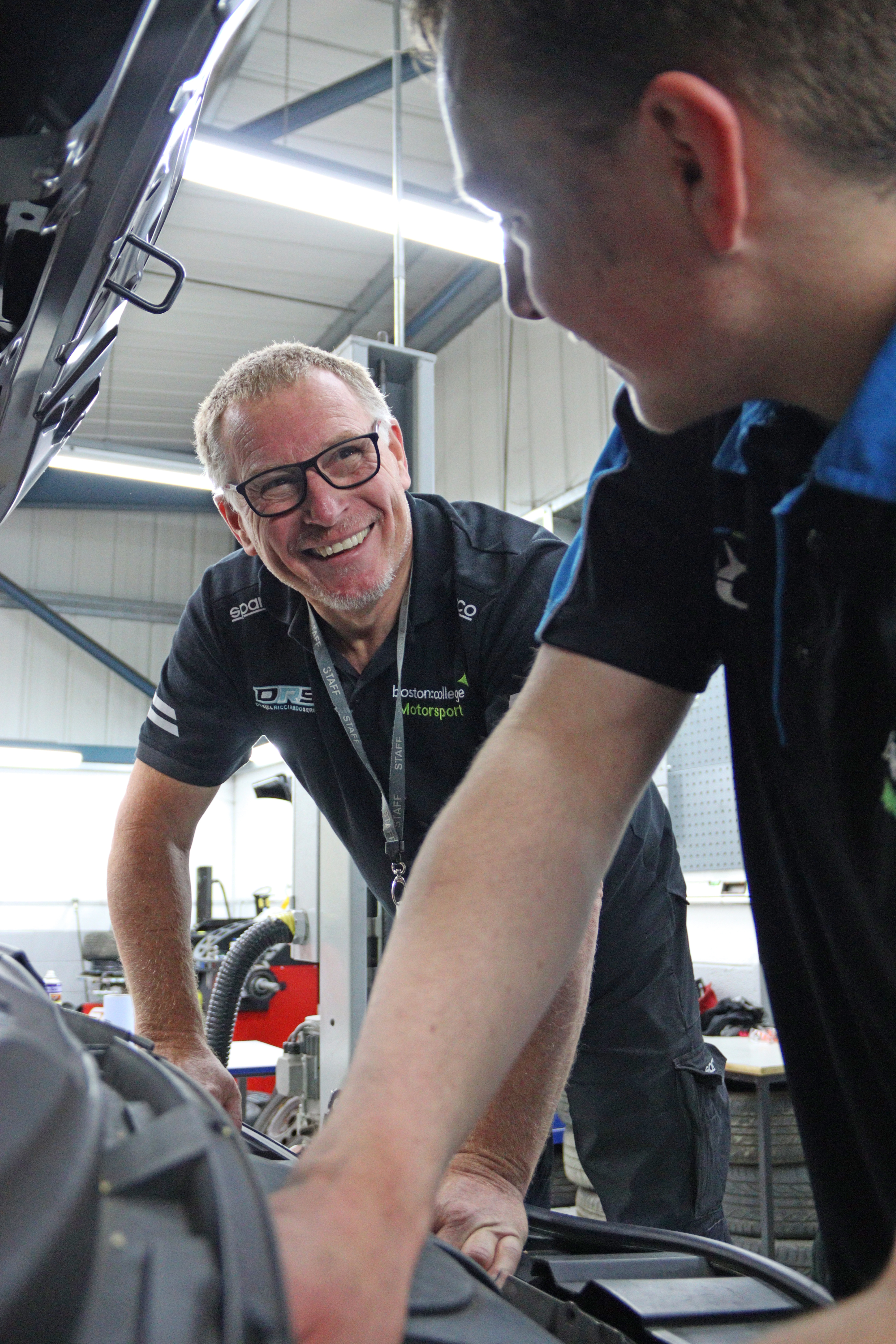 A lecturer smiles towards a student who is fixing part of an engine.