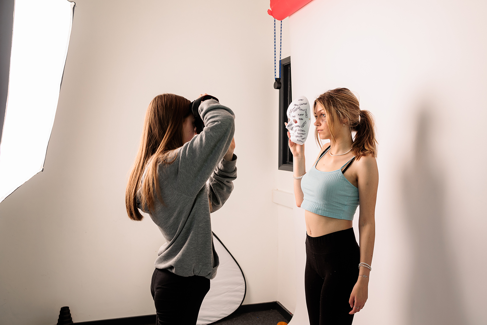 A student poses with a mask while another takes their photograph.