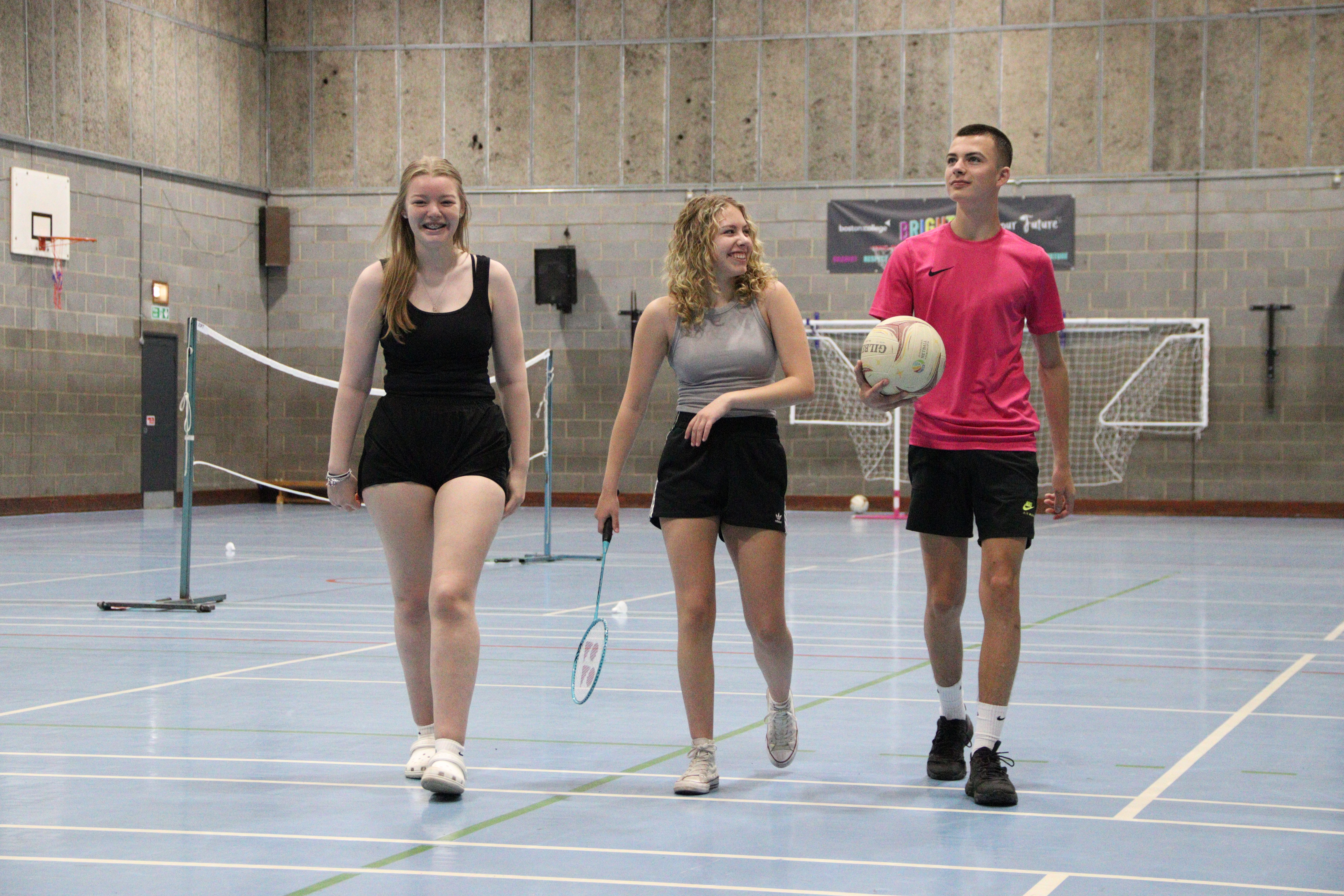 Three students in a sports hall holding sporting equipment and walking towards the camera.