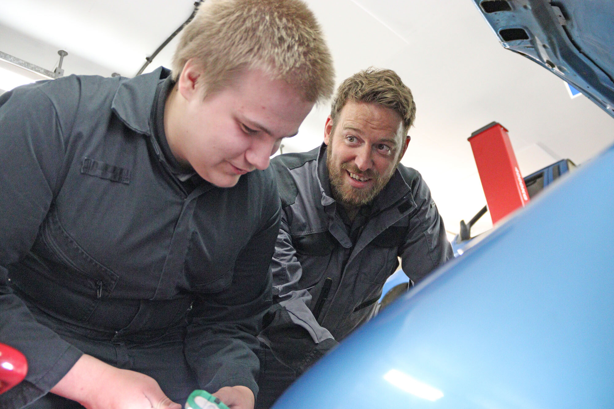 A lecturer looks on as a student fixes a car engine.
