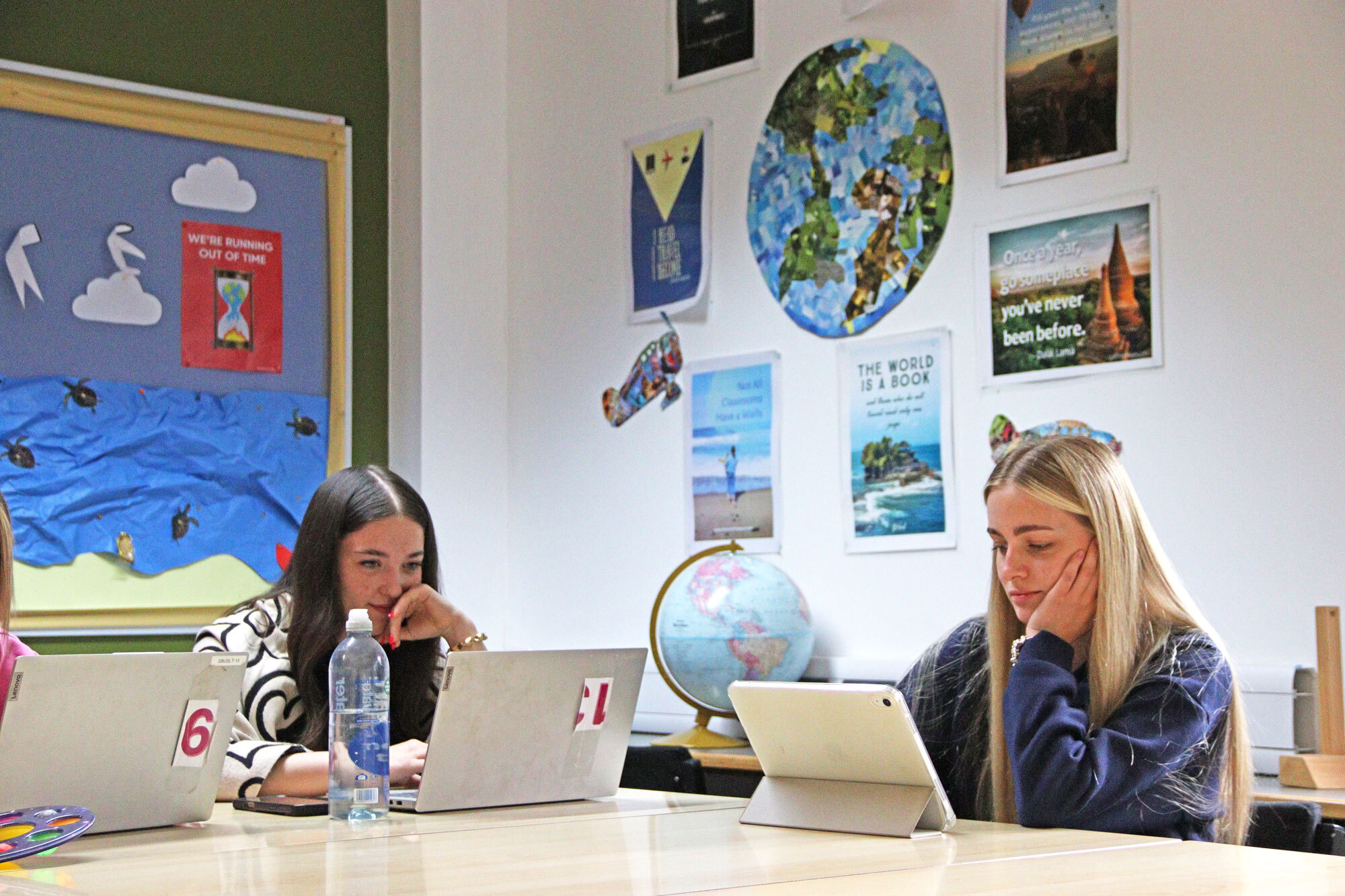 Students working on laptops at a desk with travel and tourism information on the walls.