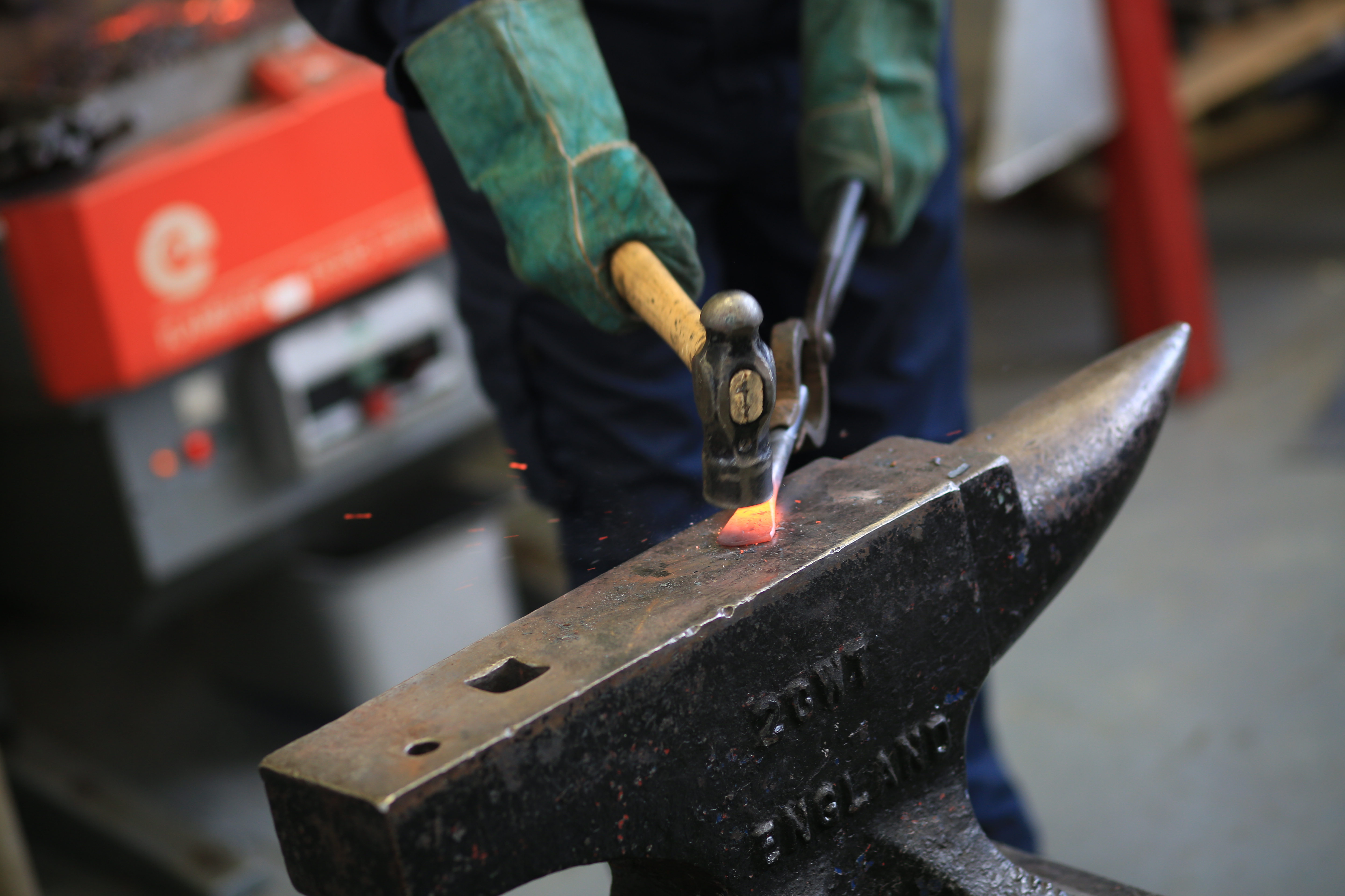 A person wearing protective gloves and clothes hammering hot metal on an anvil.