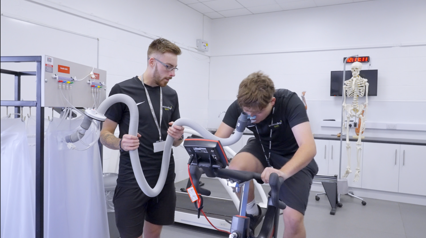 A student holds a breathing tube while another student pedals on a fitness bike.