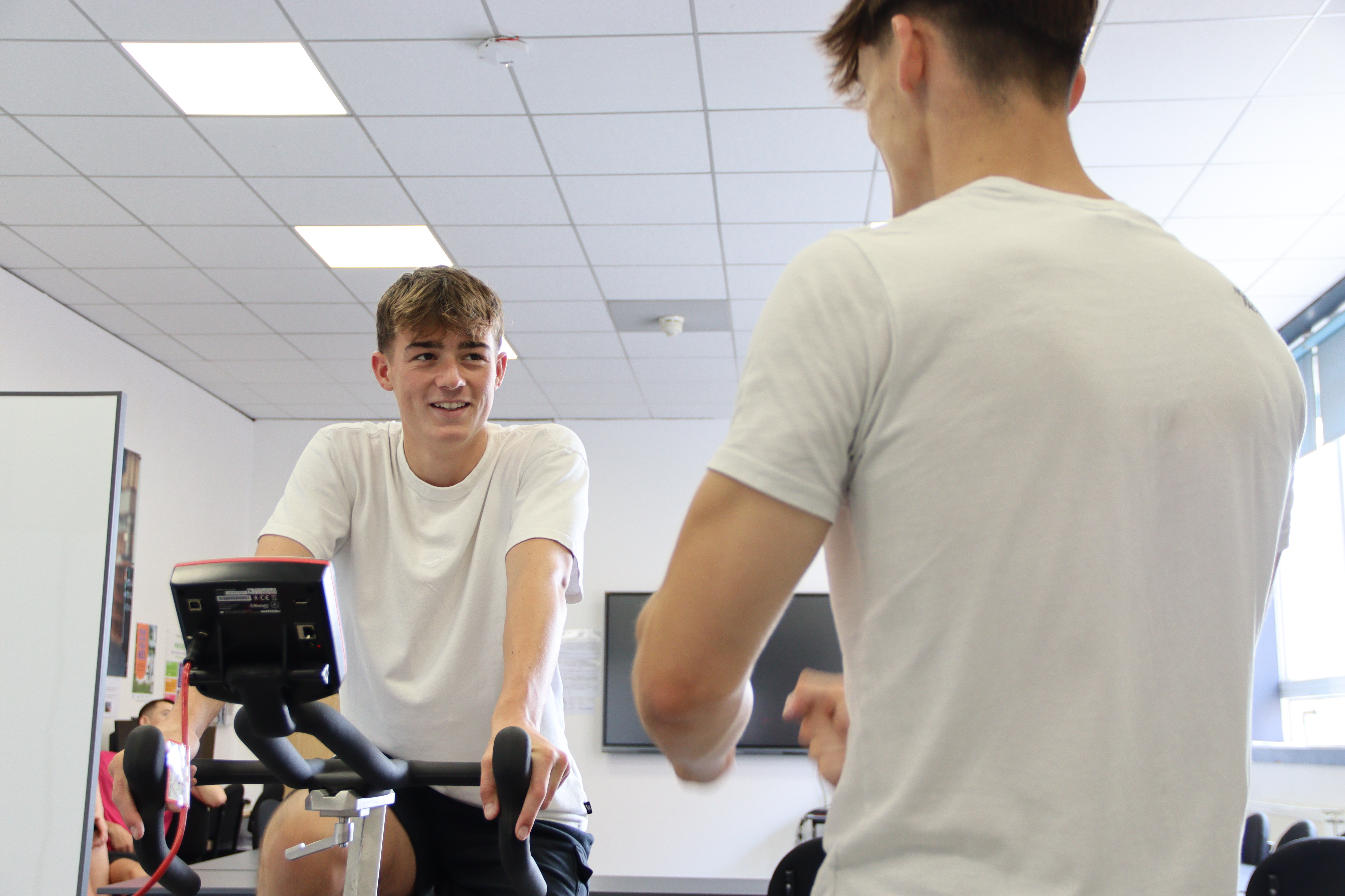 A student on an exercise bike looks up at another student supporting.