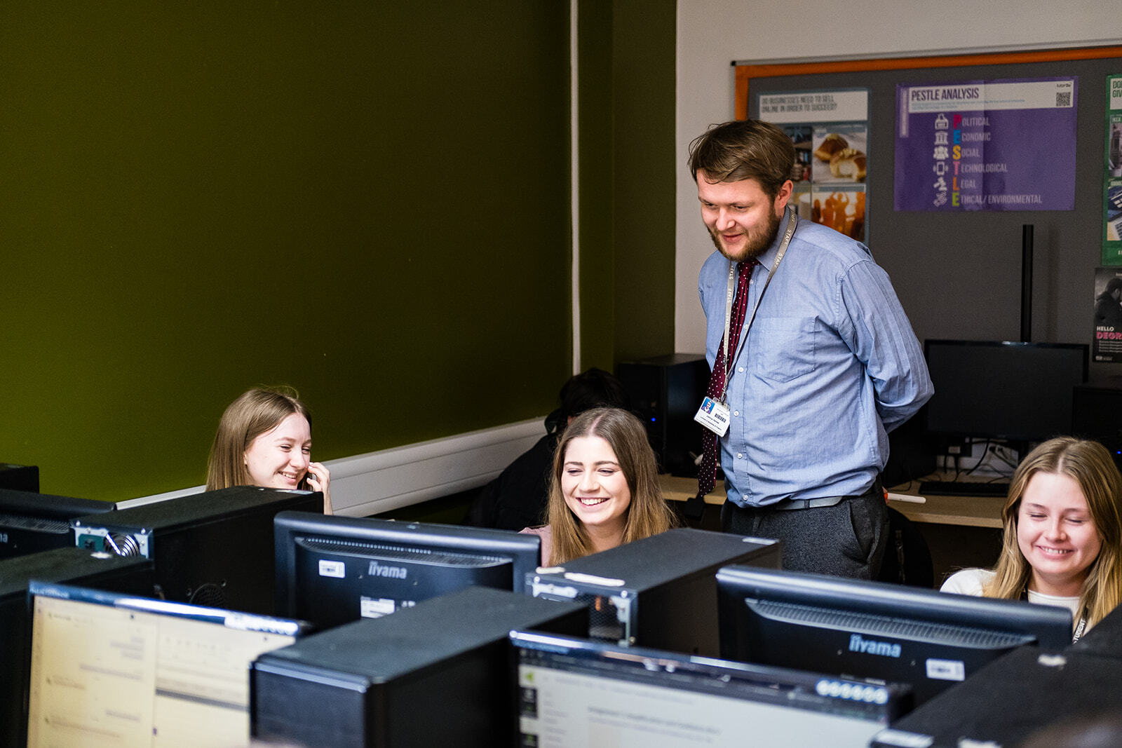 Three students sat at computers with a lecturer talking to them stood up behind them.
