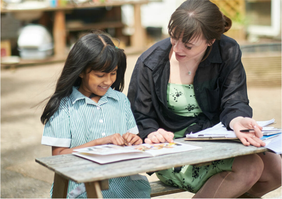 Student teacher leading a one-to-one session with a school pupil; both reading and smiling on a bench outside.