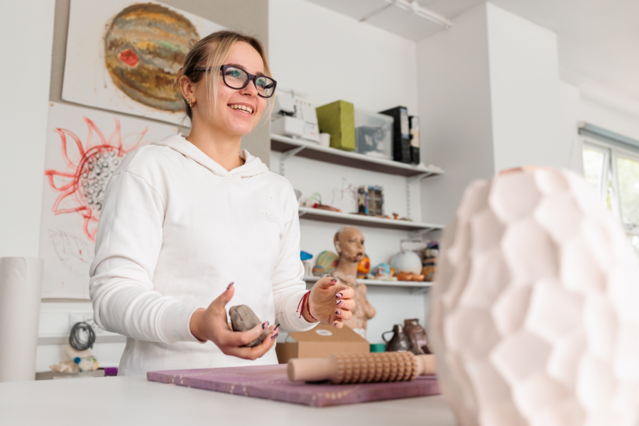 A student stood smiling in an art classroom holding modelling clay.