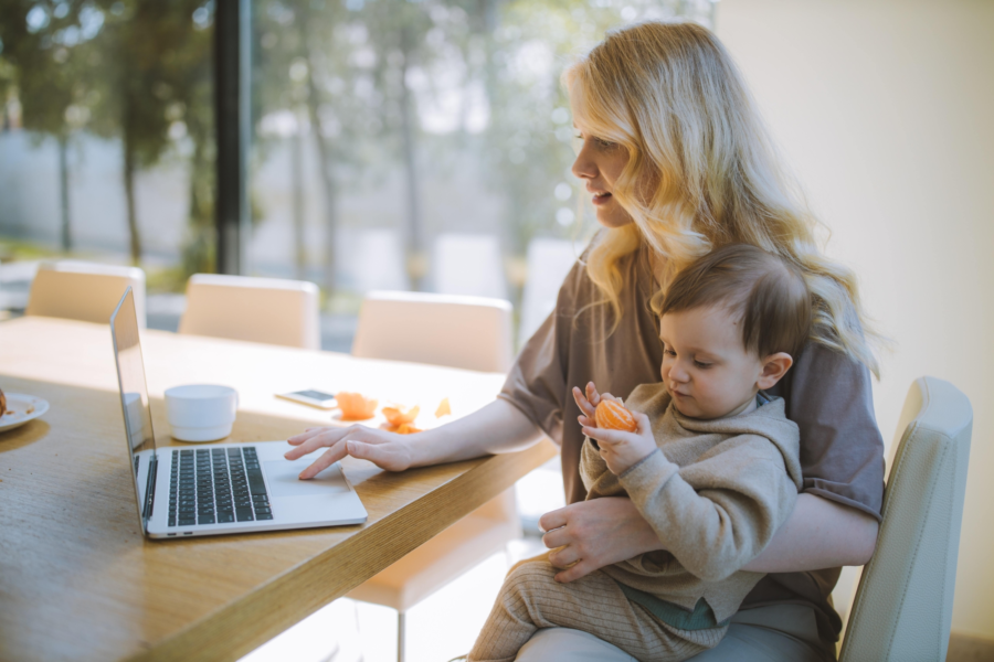 A person sat a table, working on a laptop with a baby on their knee.