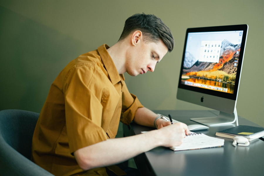 Person writing in a notebook whilst sat at a computer.