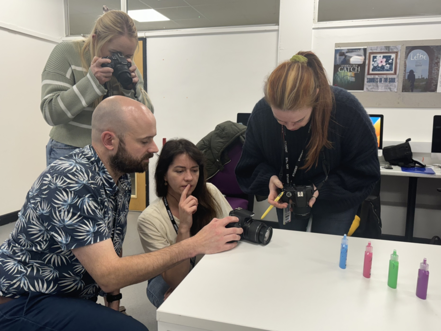 Students gather round a lecturer with a camera to discuss a task.